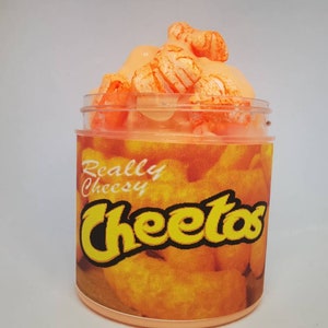Puede incluir: Un tarro de vidrio transparente lleno de slime naranja y cubierto con Cheeto puffs naranja. El tarro tiene una etiqueta que dice "Really Cheesy Cheetos".