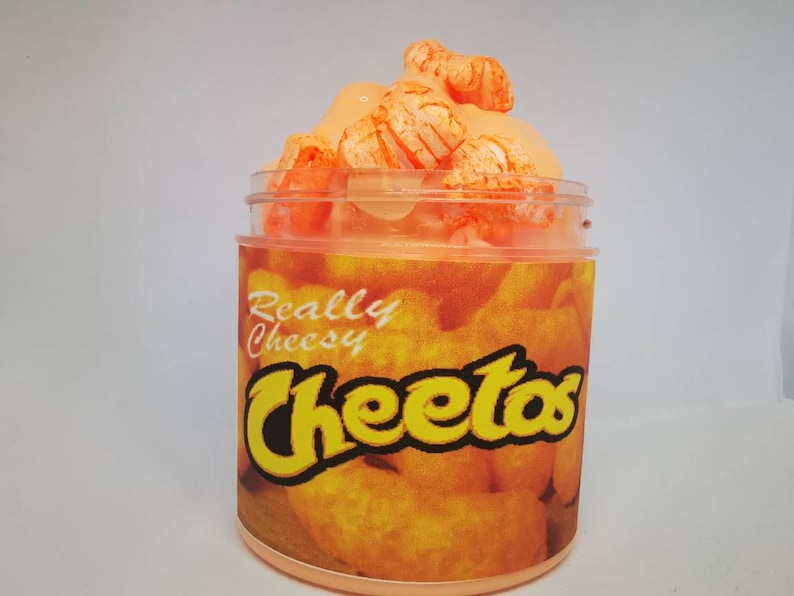 Puede incluir: Un tarro de vidrio transparente lleno de slime naranja y cubierto con Cheeto puffs naranja. El tarro tiene una etiqueta que dice "Really Cheesy Cheetos".