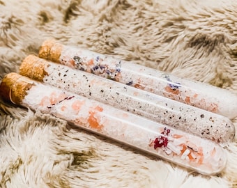 May include: Three glass test tubes filled with bath salts and dried flowers. The test tubes are on a fluffy white faux fur surface.