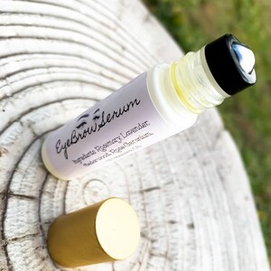 Puede incluir: Un frasco de vidrio transparente con tapa negra y tapa dorada. El frasco est&aacute; etiquetado como "Eyebrow Serum" y enumera los ingredientes: Romero, Lavanda, S&aacute;ndalo, Rosa, Geranio.