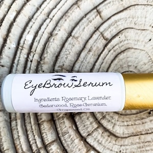 Puede incluir: Un tubo blanco y dorado con el texto "Eyebrow Serum" y una lista de ingredientes: Romero, Lavanda, Cedro, Geranio de Rosa, Aceite de Semilla de Uva.