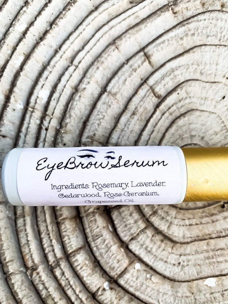 Puede incluir: Un tubo blanco y dorado con el texto "Eyebrow Serum" y una lista de ingredientes: Romero, Lavanda, Cedro, Geranio de Rosa, Aceite de Semilla de Uva.
