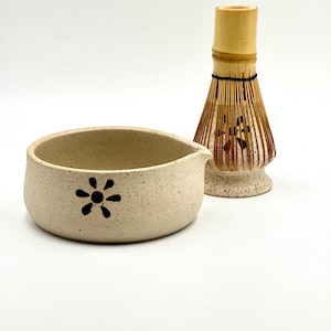 May include: A beige ceramic matcha bowl with a black floral design and a bamboo matcha whisk. The bowl has a spout for easy pouring. The whisk is made of bamboo and has a natural finish. The items are set against a white background.