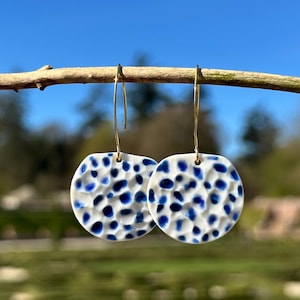 Pode incluir: Brincos redondos de cerâmica branca com superfície texturizada e um padrão de pontos azuis. Os brincos pendem de fios de orelha dourados. O fundo é uma vista desfocada de um dia ensolarado.