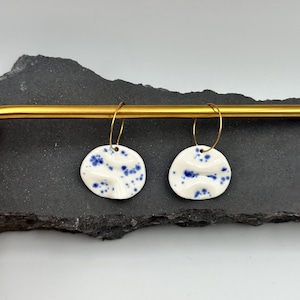 Può includere: Un paio di orecchini a cerchio con pendenti circolari in ceramica bianca caratterizzati da un motivo a macchie blu. Ogni pendente presenta un centro leggermente rientrato ed è appeso a un sottile cerchio in metallo color oro.