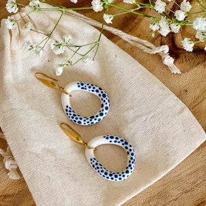 Puede incluir: Un par de pendientes de aro de cerámica blanca con un patrón de lunares azul y blanco. Los pendientes tienen ganchos dorados.