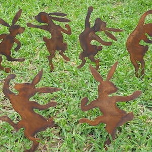 May include: Six rusty metal rabbit garden ornaments in various dancing poses are displayed on green grass. The rabbits have long ears and are in different positions, with their arms and legs outstretched.