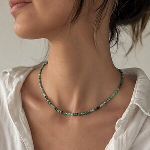 Puede incluir: Un collar de cuentas compuesto por pequeñas piedras redondas facetadas en varios tonos de verde, desde el verde menta claro hasta el verde bosque profundo, intercaladas con pequeñas cuentas de color rosa oscuro y negro, creando un patrón sutil.