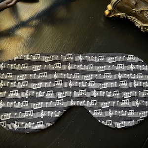 Könnte beinhalten: Schwarze Schlafmaske mit einem weißen Notenmuster. Die Maske ist aus weichem Stoff und hat ein elastisches Band.