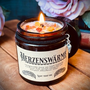 Könnte beinhalten: Ein braunes Glaskerzengefäß mit schwarzem Deckel und einem silbernen Anhänger. Die Kerze ist angezündet und hat ein hellbraunes Wachs mit kleinen roten, orangen und gelben Kristallen, die darin eingebettet sind. Das Etikett auf dem Gefäß lautet "HERZENSWÄRME" und "Handgemachte Kerze aus 100% Sojawachs mit feinem Duft, kleinen Edelsteinen und biologisch abbaubarem Glitzer" und "LIGHT YOUR FIRE".