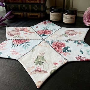 Könnte beinhalten: Ein sternförmiger Tischläufer mit einem floralen Muster in Rosa-, Grün- und Weißtönen. Der Läufer ist aus Stoff gefertigt und hat einen gesteppten Rand. In der Mitte des Läufers befindet sich eine kleine Illustration einer Person in einem grünen Kleid.