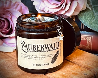 May include: A brown glass jar candle with a beige label that reads "ZAUBERWALD" and "Handgemachte Kerze aus 100% Sojawachs mit feinem Duft, kleinen Edelsteinen und biologisch abbaubarem Glitzer" and "LET THERE BE MAGIC". The candle has a silver star charm hanging from the jar.
