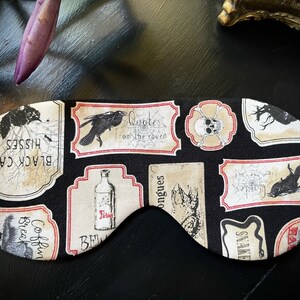 Könnte beinhalten: Schwarze Schlafmaske mit Halloween-Motiv. Die Maske ist mit verschiedenen Vintage-Etiketten verziert, die gruselige Bilder zeigen, darunter eine schwarze Katze, Totenkopf und Knochen, einen Raben und eine Zaubertrankflasche. Die Etiketten haben rote Ränder und sind auf schwarzem Hintergrund gedruckt.