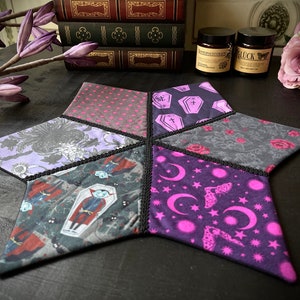 Könnte beinhalten: Ein sechszackiger sternförmiger Untersetzer mit schwarzem Rand. Jede Spitze des Sterns zeigt ein anderes Stoffdesign, darunter ein violettes Blumenmuster, ein schwarz-weißes Vampirmuster, ein rosa-violettes Mond- und Sternmuster, ein schwarz-weißes Sargmuster, ein rosa Tupfenmuster und ein schwarz-weißes Blumenmuster.