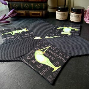 Könnte beinhalten: Vier schwarze Stoffuntersetzer mit einem weißen Streifenmuster und einem grünen Froschdesign. Jeder Untersetzer zeigt einen anderen Frosch und einen Spruch, darunter "Sei stark", "Langsam", "Lass los" und "Finde dein Gleichgewicht".