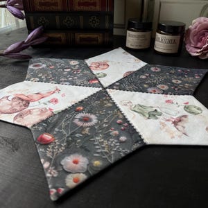 Könnte beinhalten: Ein sternförmiger Tischläufer mit Patchwork-Design, das florale Muster und Tierillustrationen zeigt. Der Läufer ist aus Stoff und hat einen dunkelgrauen Hintergrund mit weißen Nähten.