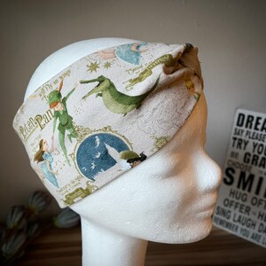 Könnte beinhalten: Ein beiges Stoffstirnband mit einem Peter Pan-Motiv. Das Design zeigt Illustrationen von Peter Pan, Wendy, einem Krokodil und anderen Figuren. Der Text enthält "Peter Pan" und "Neverland".