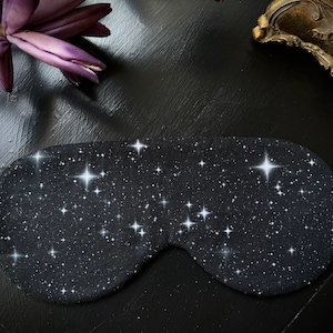 Könnte beinhalten: Eine schwarze Schlafmaske mit einem weißen Sternenhimmel-Design. Die Maske ist aus weichem Stoff gefertigt und soll Licht abhalten.