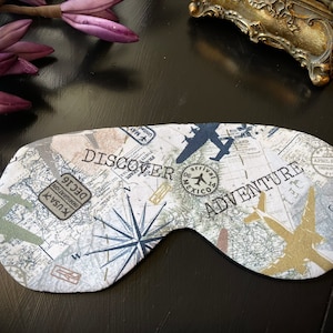 Könnte beinhalten: Eine weiße Schlafmaske mit einem Karten- und Flugzeugdesign. Die Maske trägt den Schriftzug "Discover Adventure".