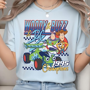 May include: A light blue t-shirt featuring a graphic print of Woody, Buzz Lightyear, and the RC car from Toy Story. The text reads "WOODY & BUZZ & RC" and "1995 Champions" in blue, yellow, and red colours, accompanied by a black and white checkered pattern.
