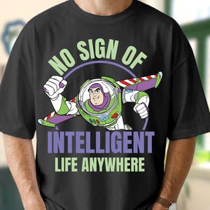 Może przedstawiać: Czarny t-shirt z grafiką Buzza Astrala w locie. Projekt zawiera tekst "NO SIGN OF INTELLIGENT LIFE ANYWHERE" w kolorze zielonym i fioletowym. Koszulka ma okrągły dekolt.