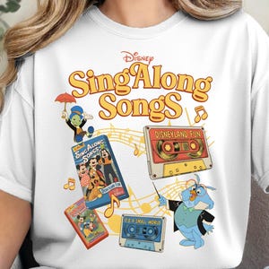 May include: White t-shirt featuring the Disney Sing Along Songs logo in yellow and orange. The shirt includes cartoon images of characters and cassette tapes with the text "Disneyland Fun" and "It's a Small World".