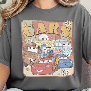 May include: A charcoal gray t-shirt featuring a graphic print with the text "CARS" in large yellow letters. The design includes illustrations of five animated vehicles, including a red race car, a brown tow truck, and a light blue van, with flowers and stars.