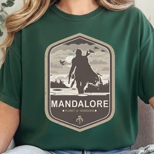 May include: A dark green t-shirt featuring a graphic print of a silhouetted warrior in a cape standing on a landscape. The design is enclosed in a hexagon with the text MANDALORE and PLANET OF WARRIORS, plus a small helmet icon at the bottom.