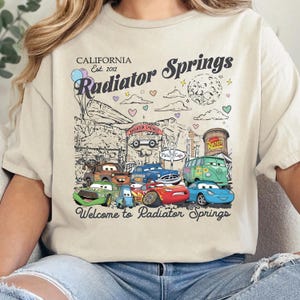 Może przedstawiać: Kremowy t-shirt z nadrukiem graficznym przedstawiającym postacie z filmu Auta. Projekt zawiera tekst California Est. 2012, Radiator Springs oraz Welcome to Radiator Springs, wraz z ilustracjami r&oacute;żnych pojazd&oacute;w.