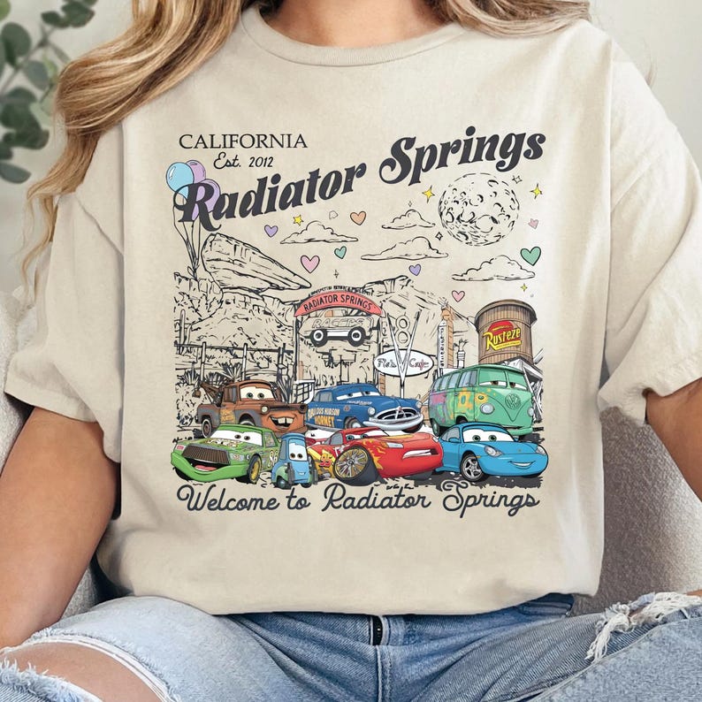 Może przedstawiać: Kremowy t-shirt z nadrukiem graficznym przedstawiającym postacie z filmu Auta. Projekt zawiera tekst California Est. 2012, Radiator Springs oraz Welcome to Radiator Springs, wraz z ilustracjami r&oacute;żnych pojazd&oacute;w.