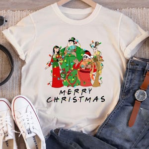 May include: Cream-colored t-shirt featuring a festive graphic of cartoon characters adorned with Christmas lights. The text "MERRY CHRISTMAS" is displayed below the characters in a stylized font. The shirt is displayed on a hanger.