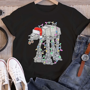 May include: Black t-shirt featuring a festive design. The graphic depicts a gray AT-AT walker wearing a Santa hat and adorned with colorful Christmas lights and ornaments. A fun, holiday-themed tee.