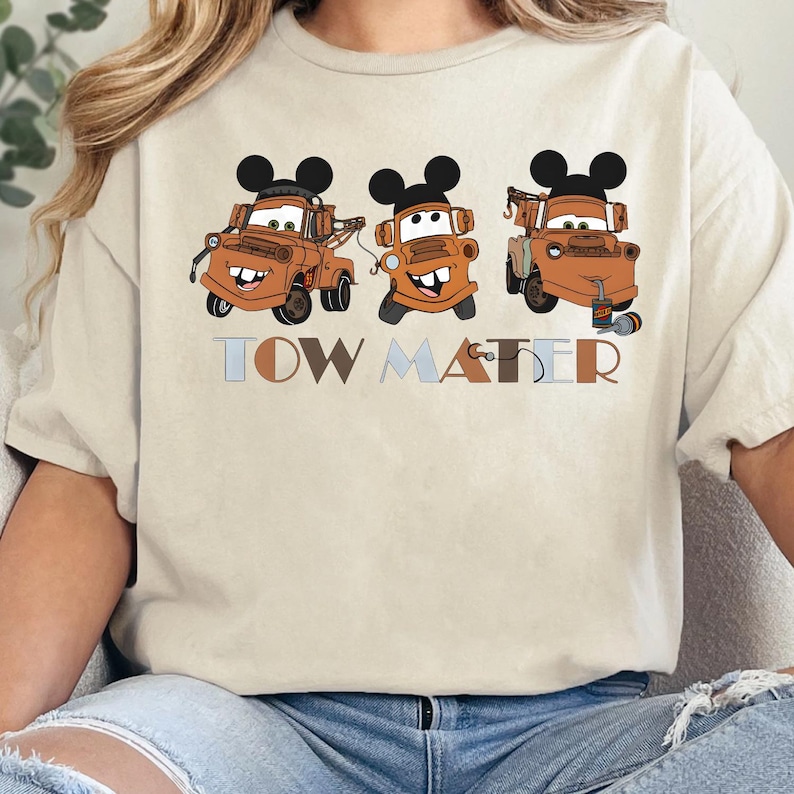 May include: A cream-colored t-shirt featuring a graphic of three brown tow trucks, each wearing black mouse ears. The trucks have smiling faces and are positioned above the text TOW MATER, which is written in a mix of light blue and brown letters.