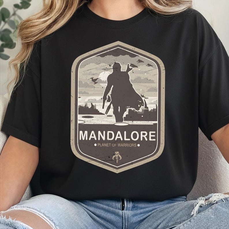 May include: A black t-shirt featuring a graphic print of a silhouetted figure in armor standing in a landscape. The design is enclosed in a beige-bordered shield shape with the text MANDALORE and PLANET OF WARRIORS printed below the figure.