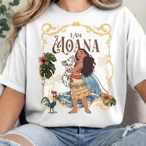 May include: A white t-shirt featuring a graphic print of a character holding a piglet, with a rooster standing nearby. The design includes blue ocean waves, green tropical leaves, pink hibiscus flowers, and the text I AM MOANA in a gold frame.