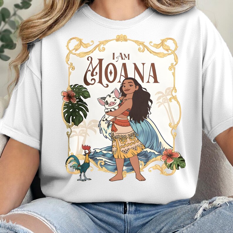 May include: A white t-shirt featuring a graphic print of a character holding a piglet, with a rooster standing nearby. The design includes blue ocean waves, green tropical leaves, pink hibiscus flowers, and the text I AM MOANA in a gold frame.
