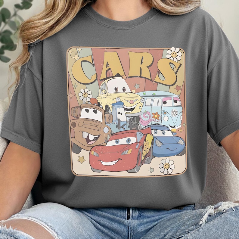 May include: A charcoal gray t-shirt featuring a graphic print with the text "CARS" in large yellow letters. The design includes illustrations of five animated vehicles, including a red race car, a brown tow truck, and a light blue van, with flowers and stars.