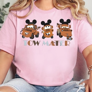 May include: A light pink t-shirt featuring a graphic of three brown tow trucks, each wearing black Mickey Mouse ears. The text "TOW MATER" is printed below the trucks in a multi-colored, block-style font with a small hook icon on the letter R.