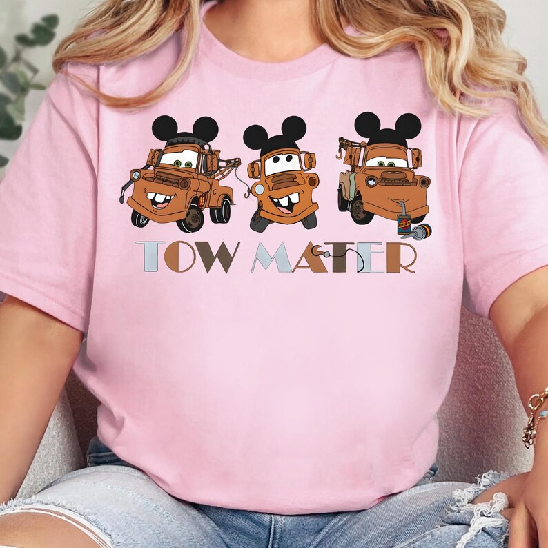 May include: A light pink t-shirt featuring a graphic of three brown tow trucks, each wearing black Mickey Mouse ears. The text "TOW MATER" is printed below the trucks in a multi-colored, block-style font with a small hook icon on the letter R.