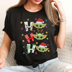 A woman wearing a black t-shirt with a graphic design featuring three cartoon characters, including a baby alien wearing a Santa hat.