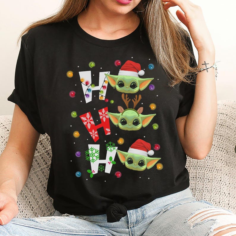 A woman wearing a black t-shirt with a graphic design featuring three cartoon characters, including a baby alien wearing a Santa hat.