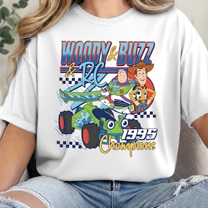 May include: A white t-shirt featuring a graphic print of Woody and Buzz Lightyear standing next to the RC car. The design includes the text "WOODY & BUZZ & RC" and "1995 Champions" in blue, red, and yellow colours with a checkered flag pattern.