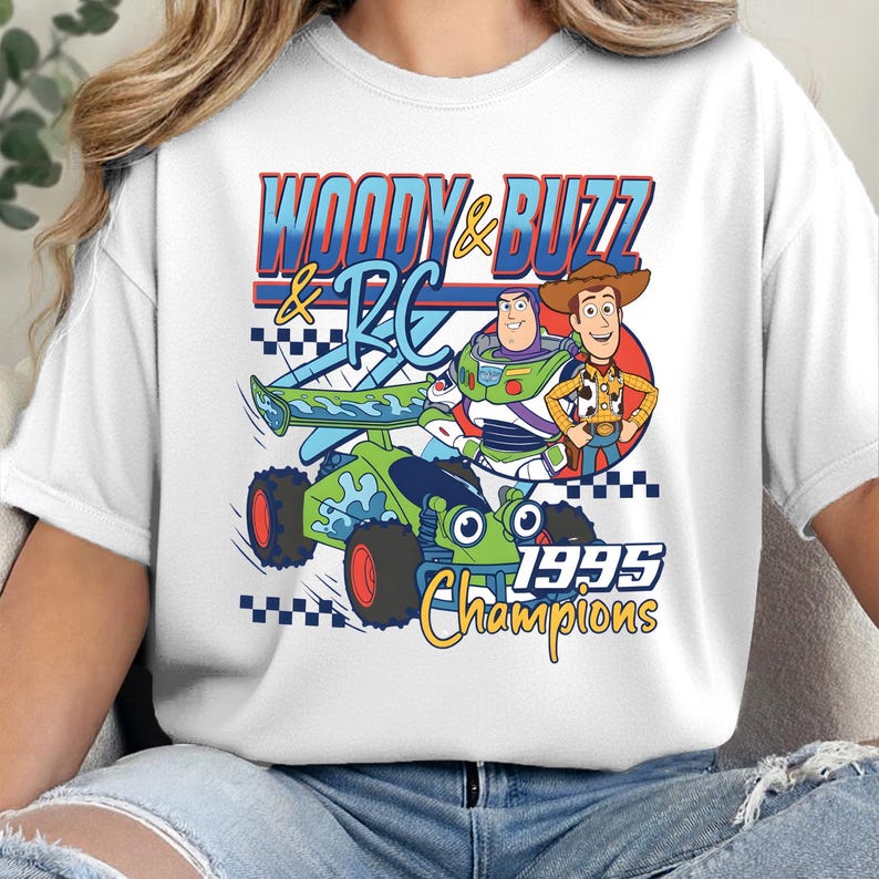 May include: A white t-shirt featuring a graphic print of Woody and Buzz Lightyear standing next to the RC car. The design includes the text "WOODY & BUZZ & RC" and "1995 Champions" in blue, red, and yellow colours with a checkered flag pattern.