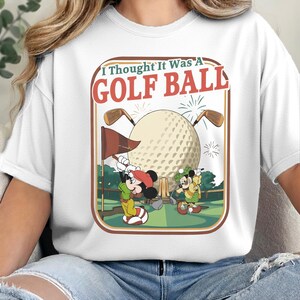 Op de afbeelding: Een wit t-shirt met een grafische print van twee tekenfilmmuizen op een golfbaan. De tekst "I Thought It Was A GOLF BALL" staat boven een grote golfbal, twee golfclubs, een vlag en groene landschapselementen in een retro kader.