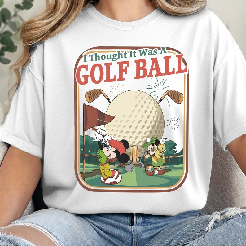 Op de afbeelding: Een wit t-shirt met een grafische print van twee tekenfilmmuizen op een golfbaan. De tekst "I Thought It Was A GOLF BALL" staat boven een grote golfbal, twee golfclubs, een vlag en groene landschapselementen in een retro kader.