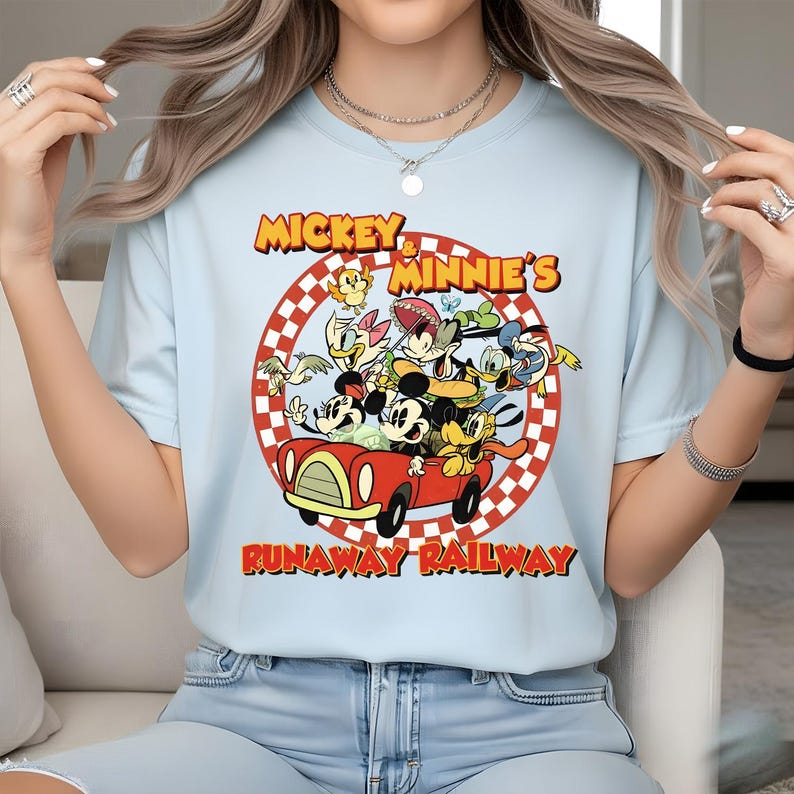 May include: Light blue t-shirt featuring a colorful graphic of Mickey and Minnie Mouse with other Disney characters in a red car. The text "Mickey & Minnie's Runaway Railway" is printed above and below the image.