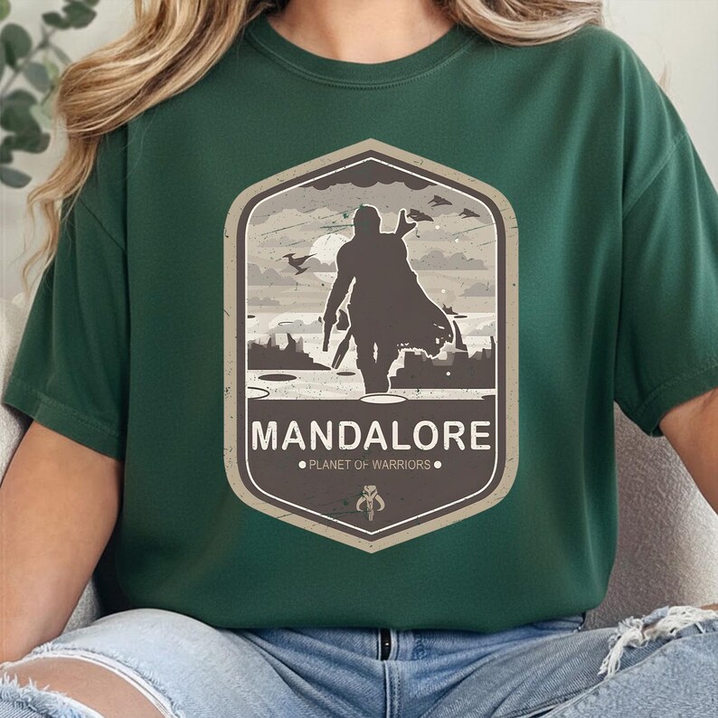 May include: A dark green t-shirt featuring a graphic print of a silhouetted warrior in a cape standing on a landscape. The design is enclosed in a hexagon with the text MANDALORE and PLANET OF WARRIORS, plus a small helmet icon at the bottom.