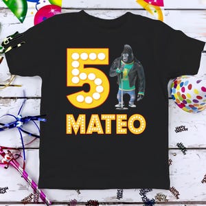 May include: Black t-shirt featuring a yellow number 5 with white dot accents and the name "MATEO" in yellow. A cartoon gorilla wearing a black jacket and holding a microphone is on the right. Party-themed apparel.