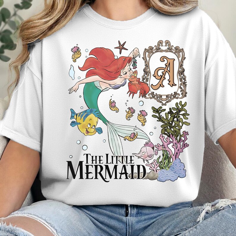 Puede incluir: Una camiseta blanca con un gr&aacute;fico de Ariel de La Sirenita, un cangrejo, un pez amarillo y un pez rosa. El dise&ntilde;o incluye un marco decorativo con la letra A, plantas marinas y el texto The Little Mermaid en letras de color negro.