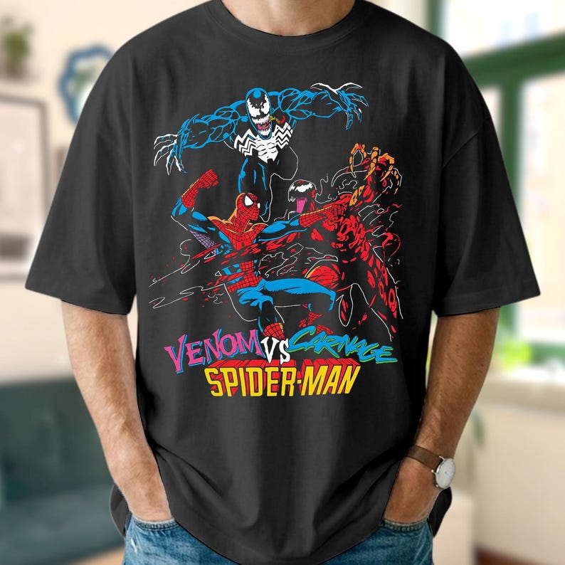 May include: Black t-shirt with a comic book style graphic of Venom, Carnage, and Spider-Man. The text "Venom vs. Carnage Spider-Man" is in a multi-coloured font. The design depicts the characters in dynamic action poses, with bold lines and bright colours.